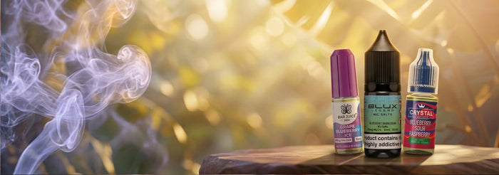 Three small e-liquid bottles from the brand M-LUX stand on a wooden surface against a warm, sunlit bokeh background with purple smoke drifting from the left. The bottles are purple, black, and blue, with labels for flavors like Mango Custard, Strawberry Milk, and Blue Raspberry Lemonade.